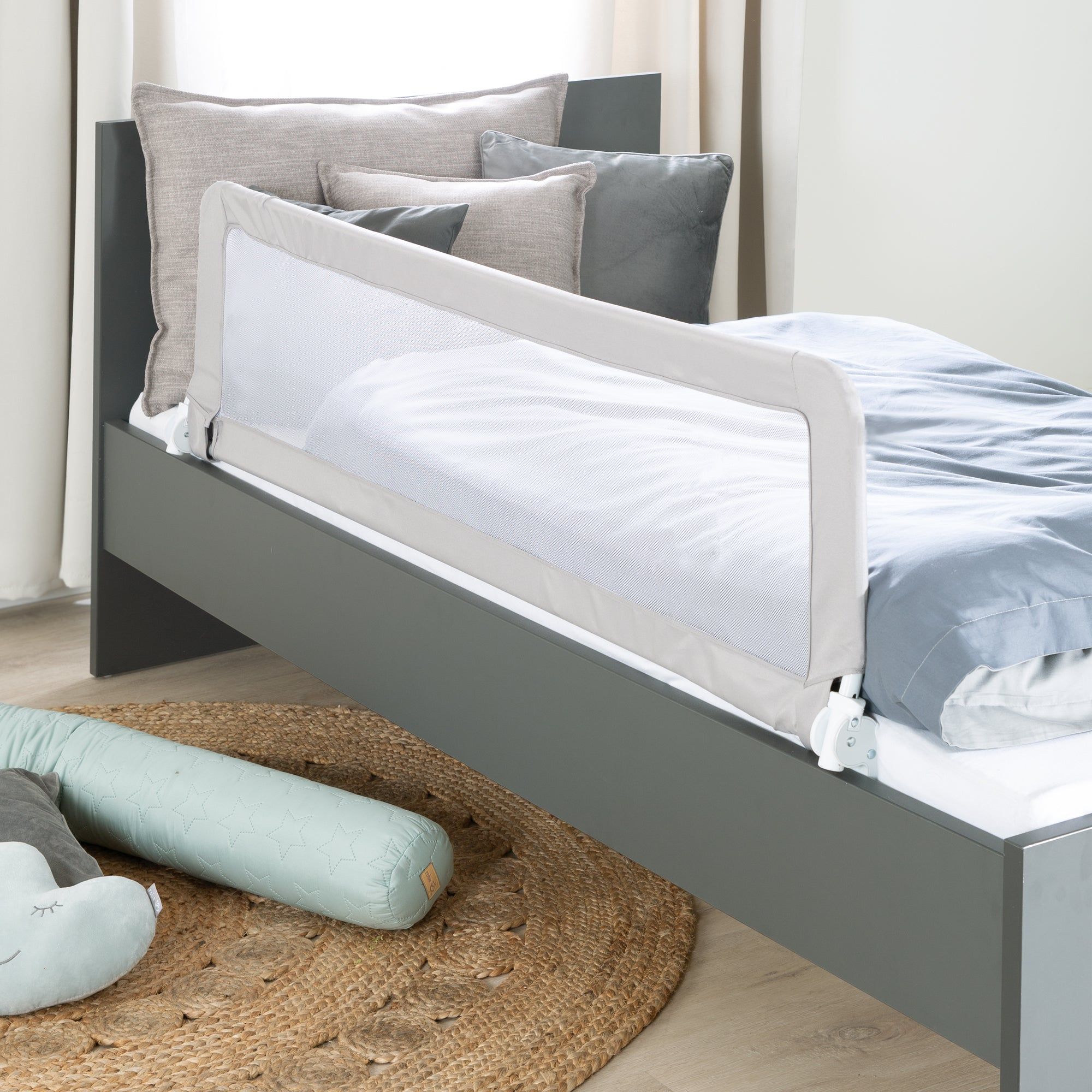 Bettgitter, weißes Netz, grauer Rahmen, am Bett befestigt. Auf dem Bett liegen Kissen und Decke.