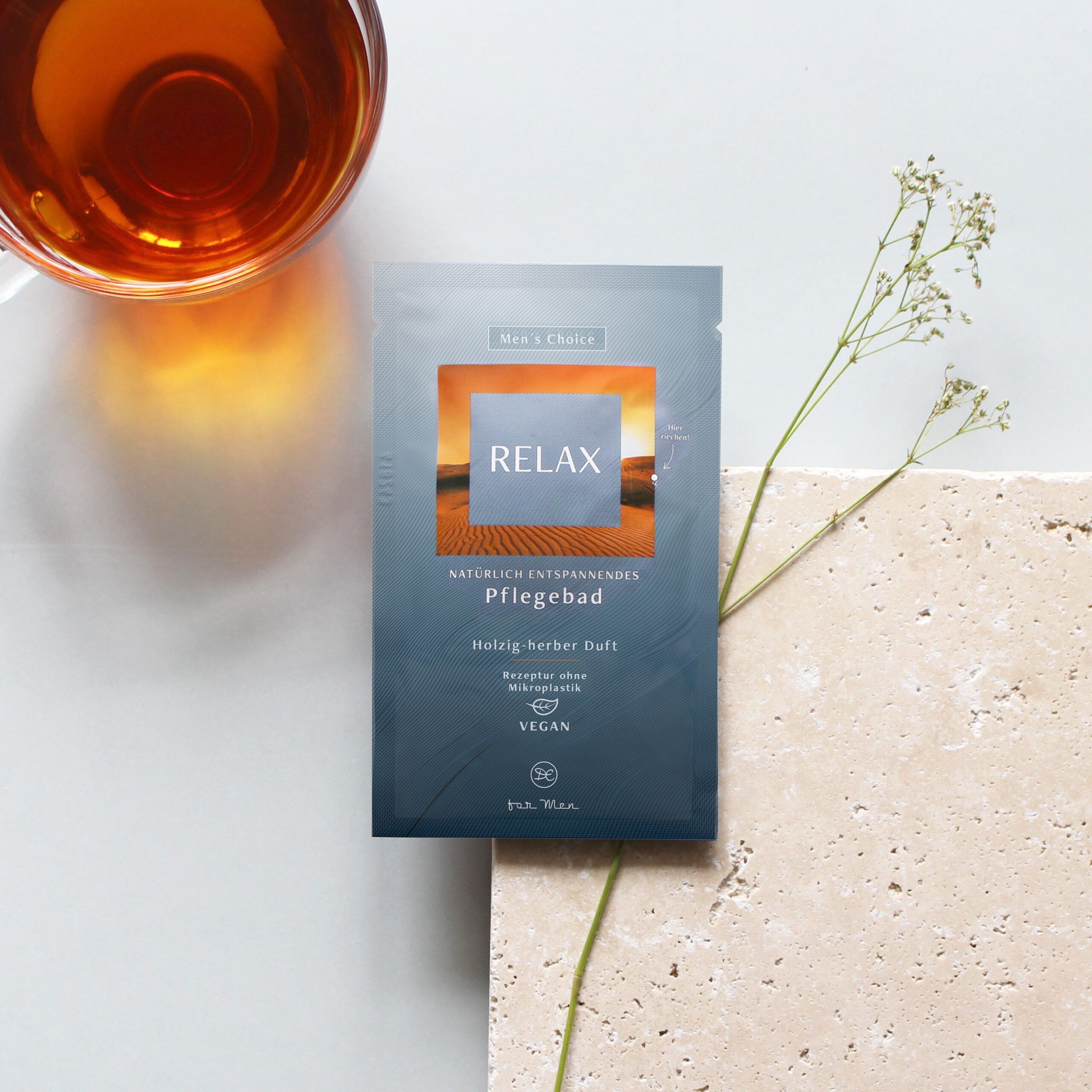 Einzelpackung RELAX auf heller Oberfläche. Neben Packung: Glas mit Flüssigkeit, Pflanze. Text: Pflegebad, holzig-herber Duft, vegan.