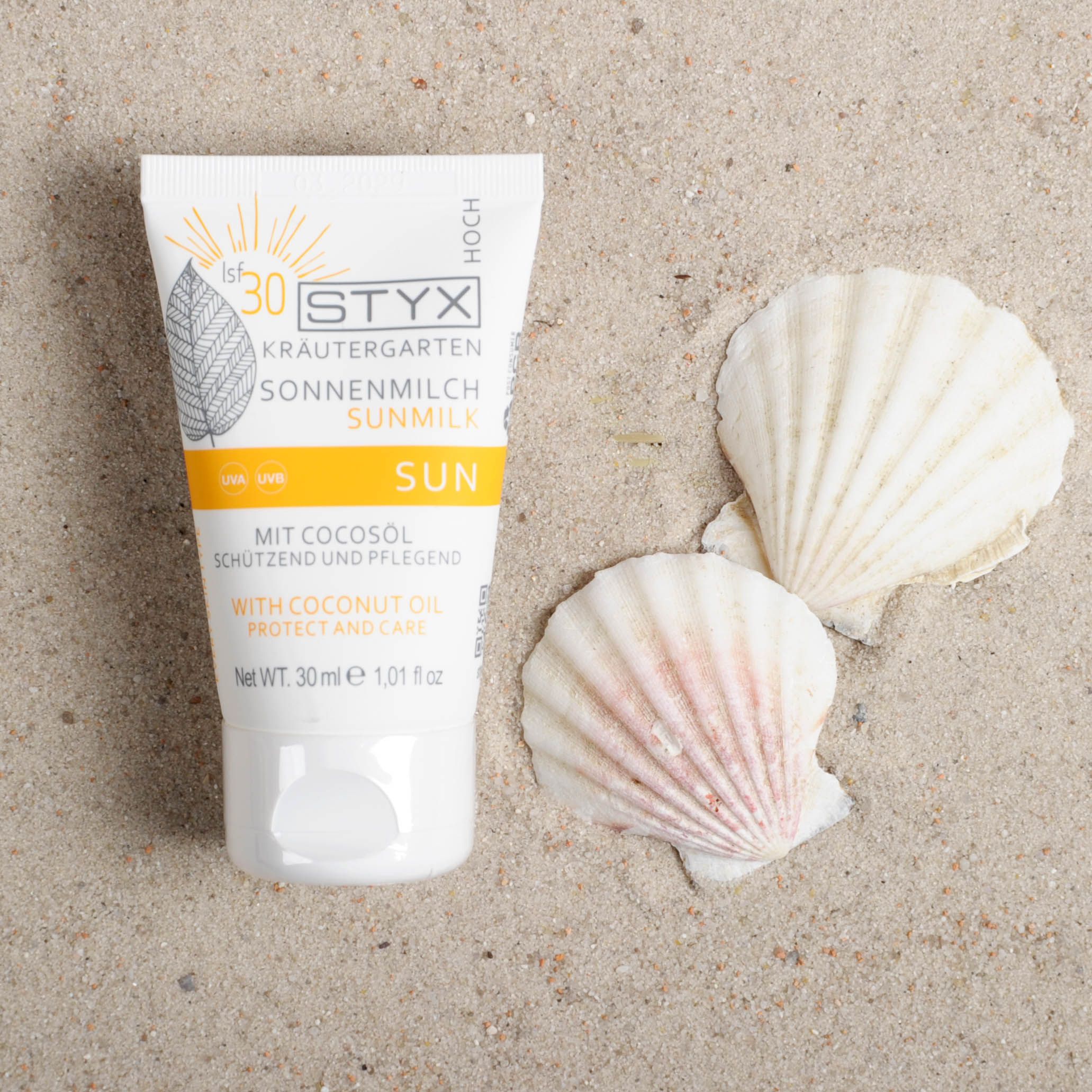 STYX Sonnenmilch LSF 30 Tube auf Sand. Weiß, mit gelben und grünen Akzenten. Text: Sonnenmilch, Sun, mit Cocosöl. 30 ml. Muscheln.