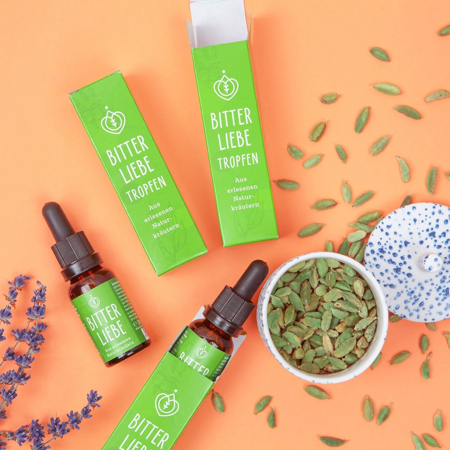 Zwei Flaschen und zwei Schachteln BitterLiebe-Tropfen. Daneben Kardamom und eine Schale.