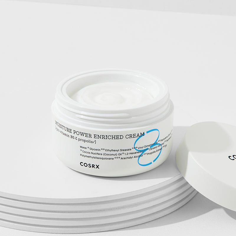 Geöffnete weiße Creme-Dose auf weißem Podest. Aufschrift: MOISTURE POWER ENRICHED CREAM, COSRX. Deckel daneben.
