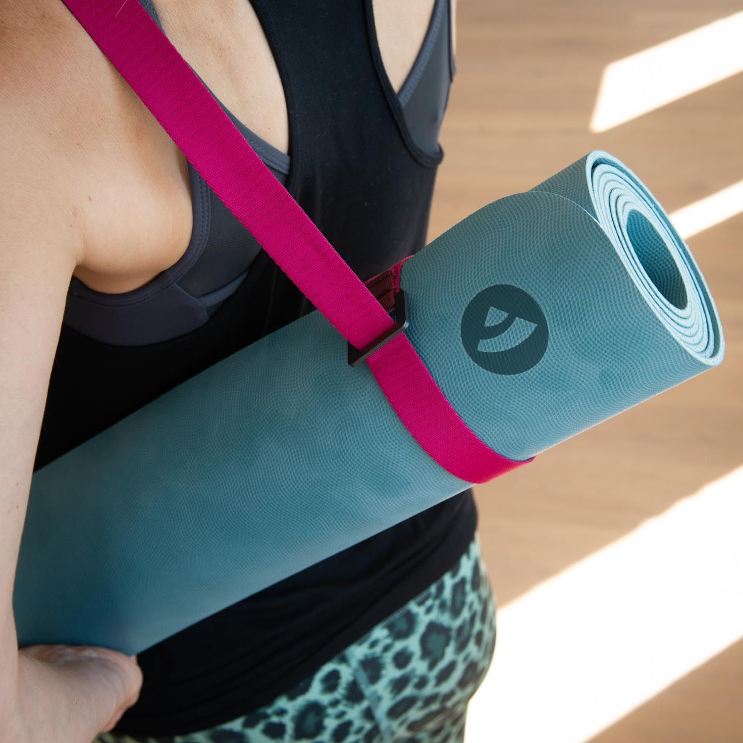 Person trägt gerollte Yogamatte mit Tragegurt. Gurt pink. Matte blau. Logo sichtbar.