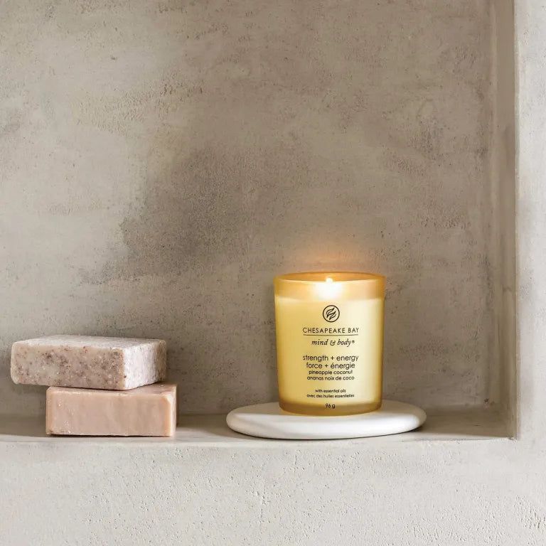 Gelbe Kerze Chesapeake Bay mind & body, Stärke + Energie, Ananas Kokosnuss, auf weißem Untersetzer. Zwei Seifen daneben.