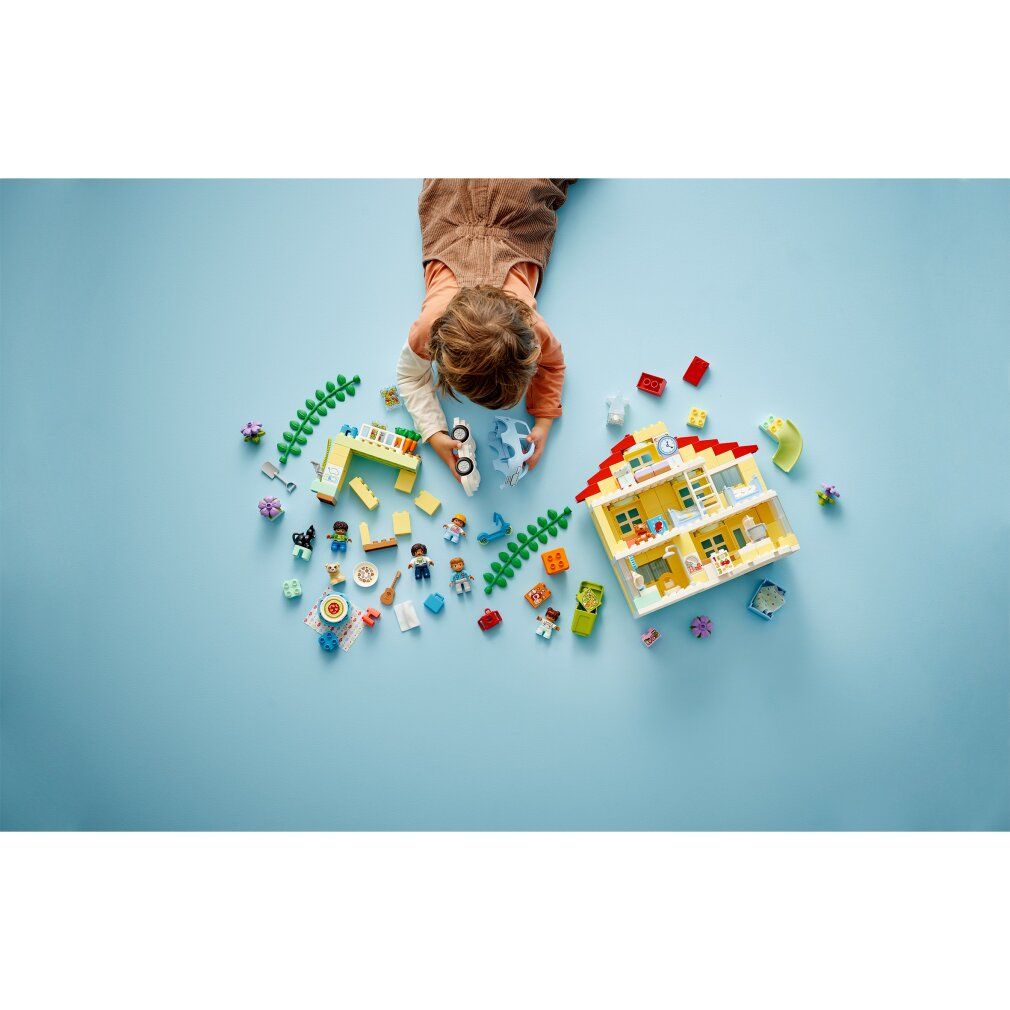 Kind spielt mit LEGO Duplo 3-in-1-Familienhaus. Gelbes Haus, Figuren, Auto, Rutsche und Zubehör auf blauem Hintergrund.