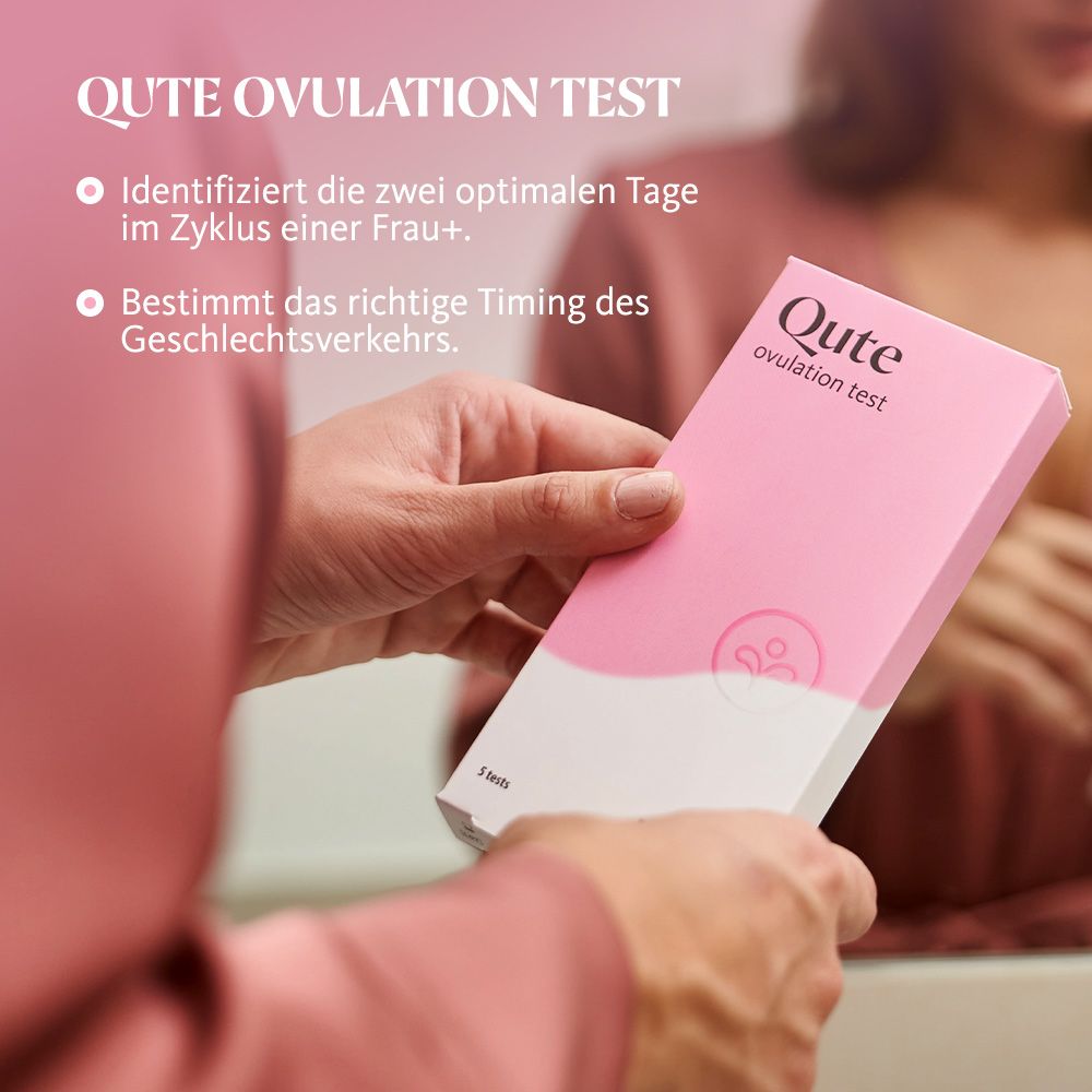 Eine Frau hält eine Qute Ovulationstest-Packung. Aufschrift: Qute Ovulationstest. Rosa Verpackung mit weißem Text.