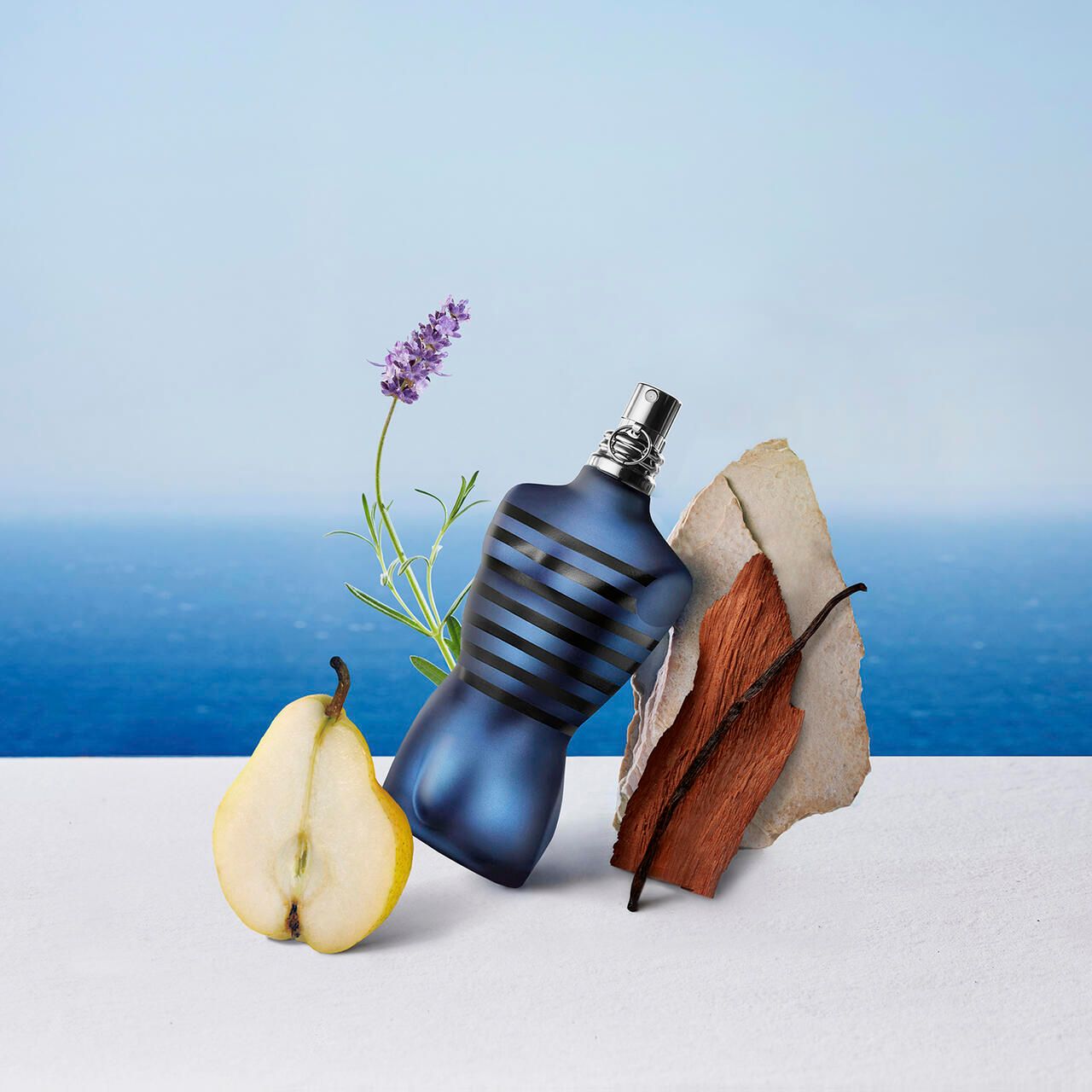 Blauer Flakon in Torsoform, umgeben von Lavendel, Birne, Holz und Stein. Hintergrund: Meer.