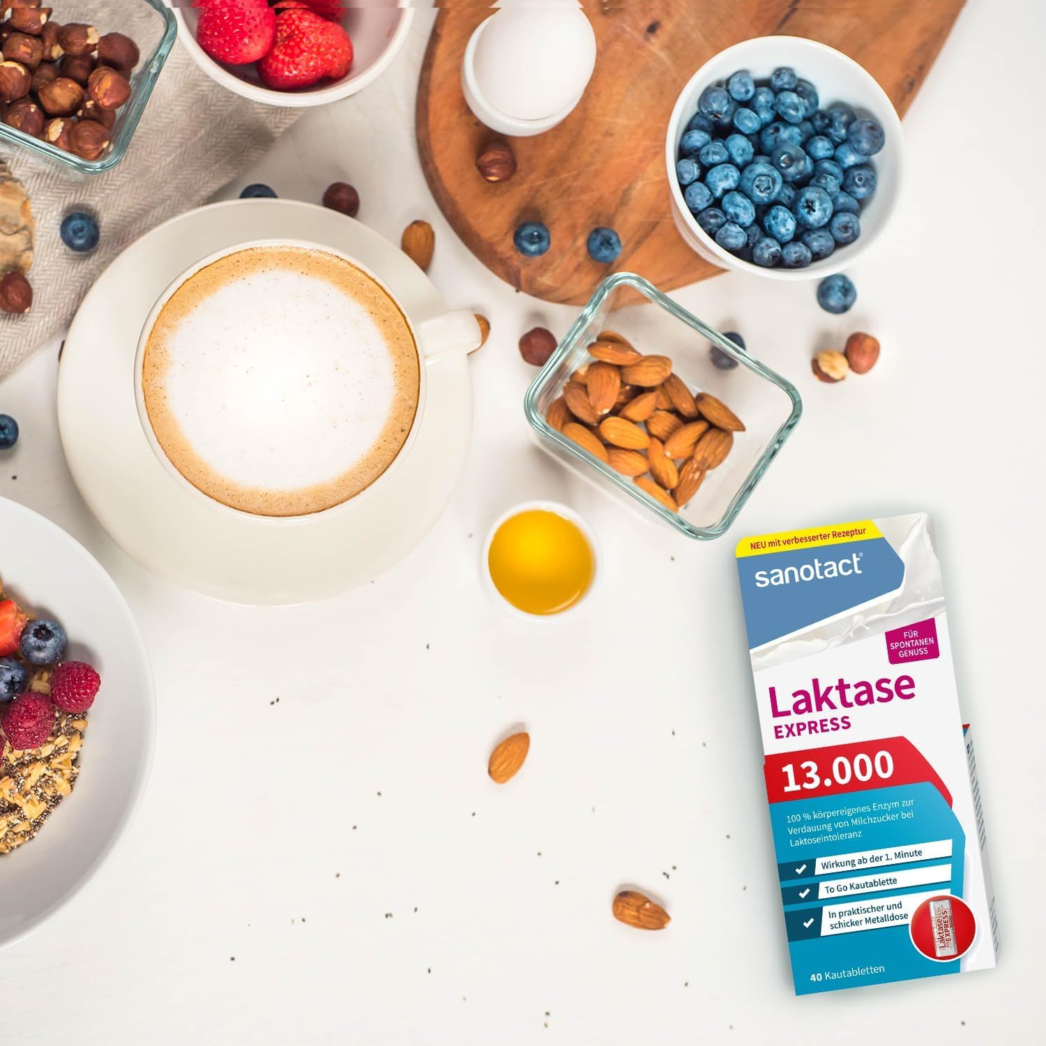 sanotact Laktase 13.000 EXPRESS Packung auf weißem Tisch. Umgeben von Lebensmitteln: Kaffee, Beeren, Nüsse, Eier, Mandeln.