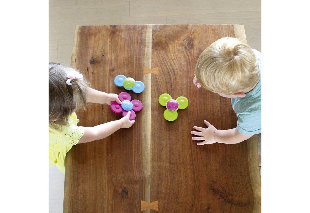 Zwei Kinder spielen mit bunten Spinnern auf einem Holztisch. Die Spinner sind in Grün, Pink und Blau.