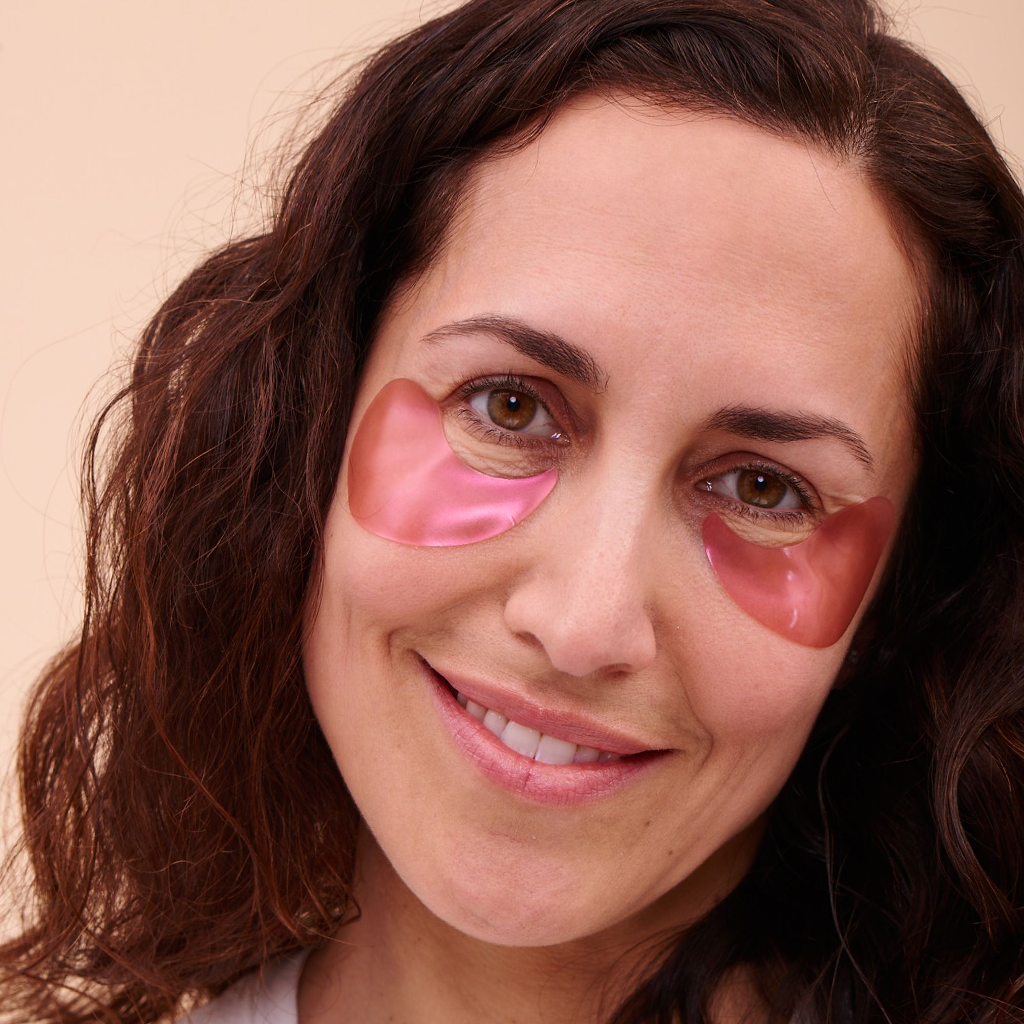 Frau mit Augenpads. Rosa Pads unter den Augen. Frau lächelt. Helle Haut, braune Augen, braune Haare.