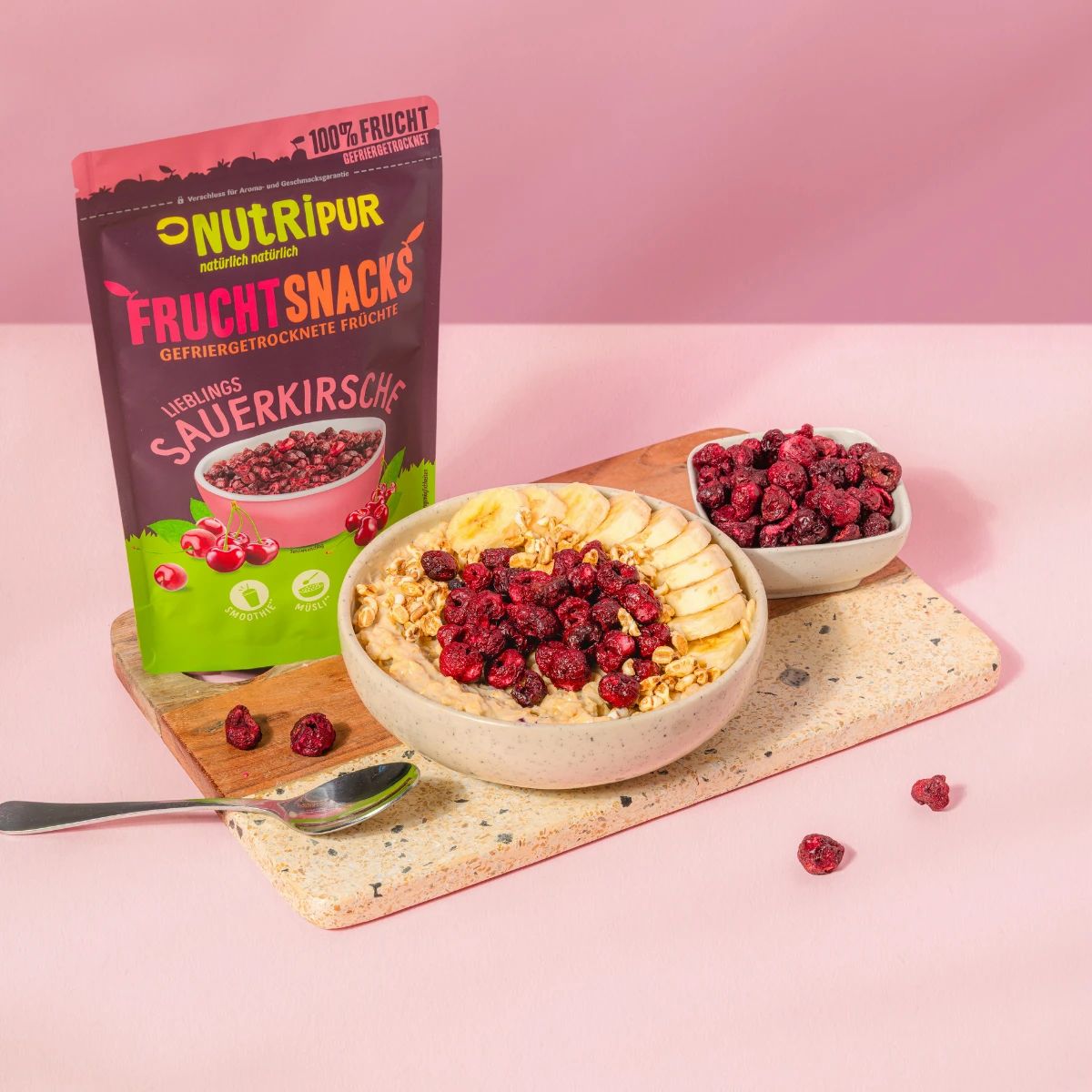 Schale mit Müsli, Bananenscheiben und gefriergetrockneten Sauerkirschen. Daneben Beutel mit Sauerkirschen. Rosa Hintergrund.