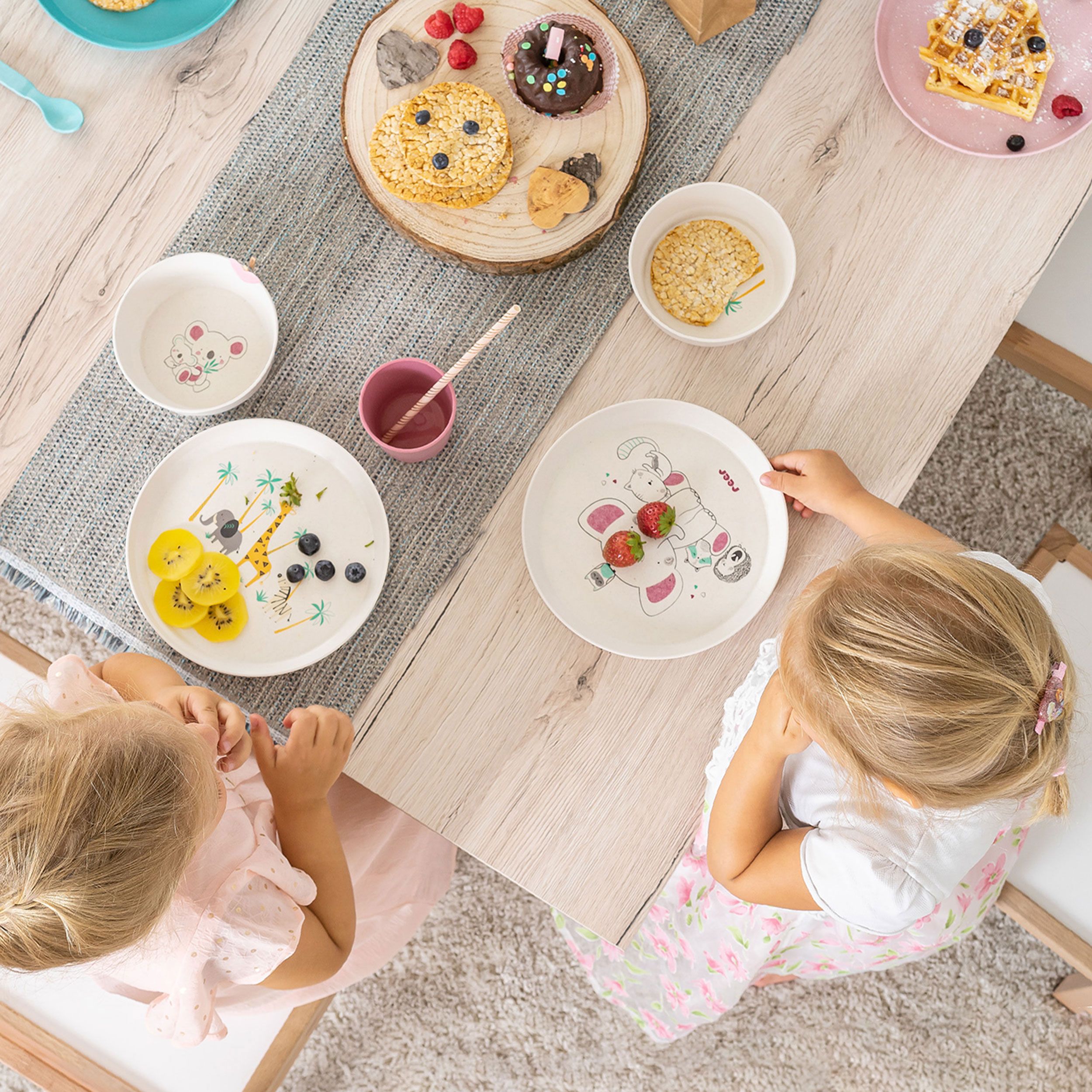 Kindergeschirr-Set mit Tellern, Schalen, Bechern und Besteck. Auf den Tellern sind Tiermotive abgebildet. Kinder sitzen am Tisch.