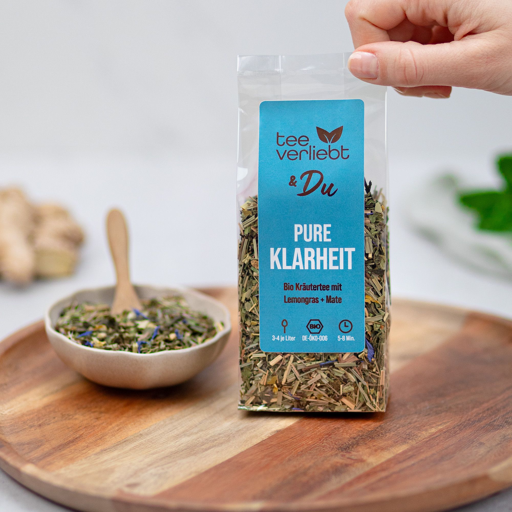 Hand hält Teebeutel. Auf Holztablett: Schale mit Kräutern, Holzlöffel. Teebeutel mit blauem Etikett.