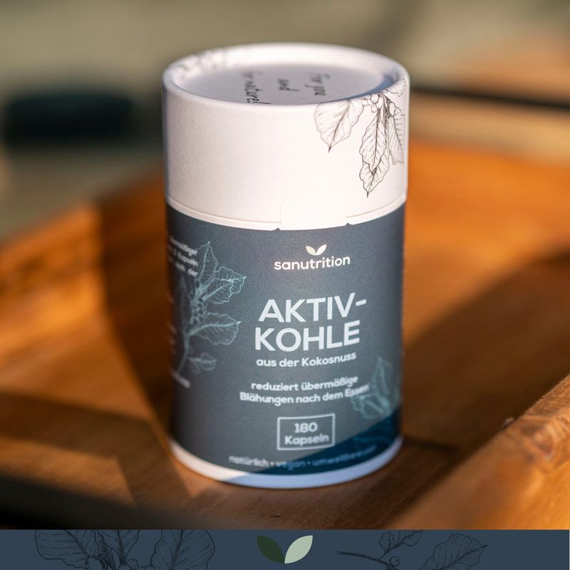 Zylindrische Dose Sanutrition Aktivkohle auf Holzuntergrund. Aufschrift: Aktivkohle aus Kokosnuss, 180 Kapseln. Reduziert Blähungen.
