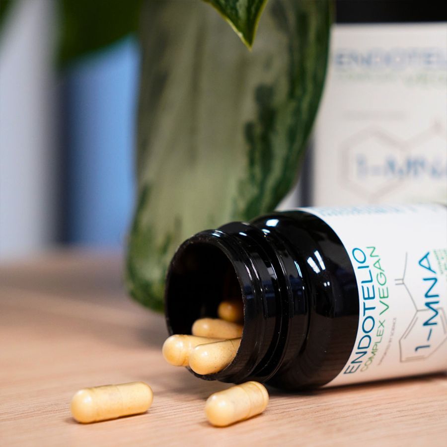 Braune Flasche mit Kapseln, die auf eine Holzoberfläche gefallen sind. Aufschrift: Endotelio Complex Vegan, 1-MNA. Einige Kapseln liegen daneben.