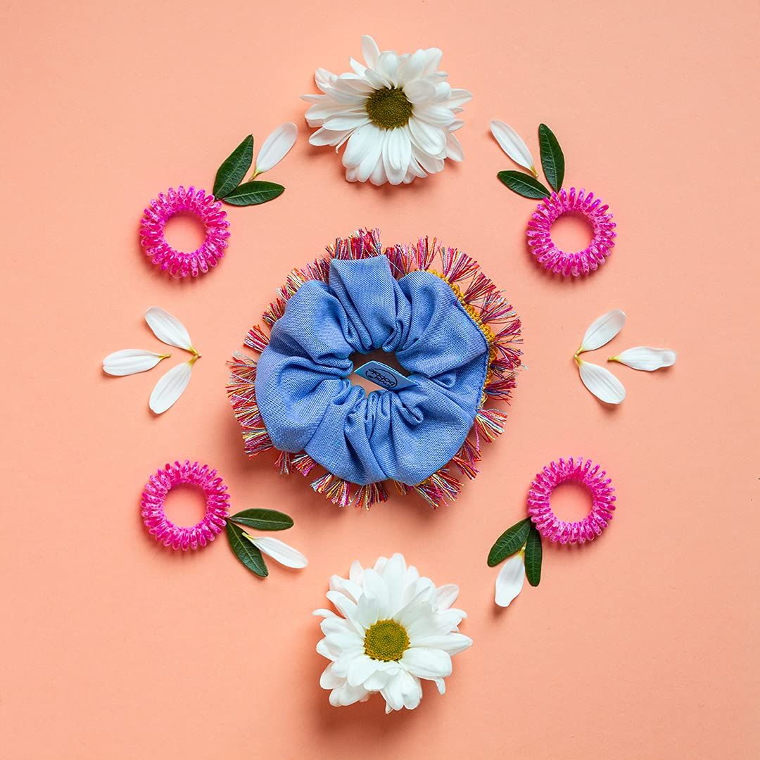 Blaues Scrunchie mit Fransen, umgeben von rosa Scrunchies, weißen Gänseblümchen und Blättern auf pfirsichfarbenem Hintergrund.