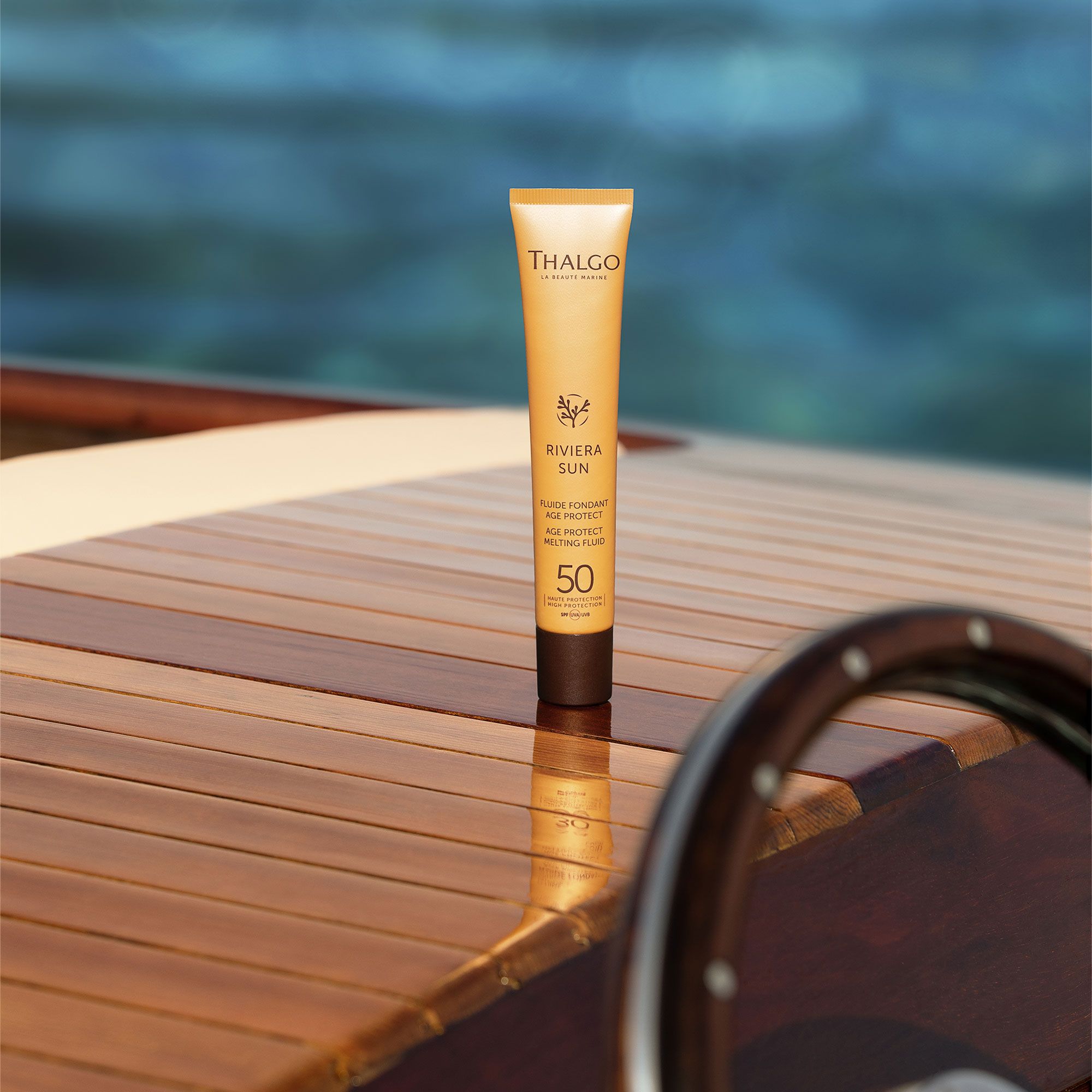 Gelbe Tube THALGO Riviera Sun auf Holzoberfläche. Enthält Age Protect Melting Fluid LSF 50. Im Hintergrund Meer und Steuerrad.
