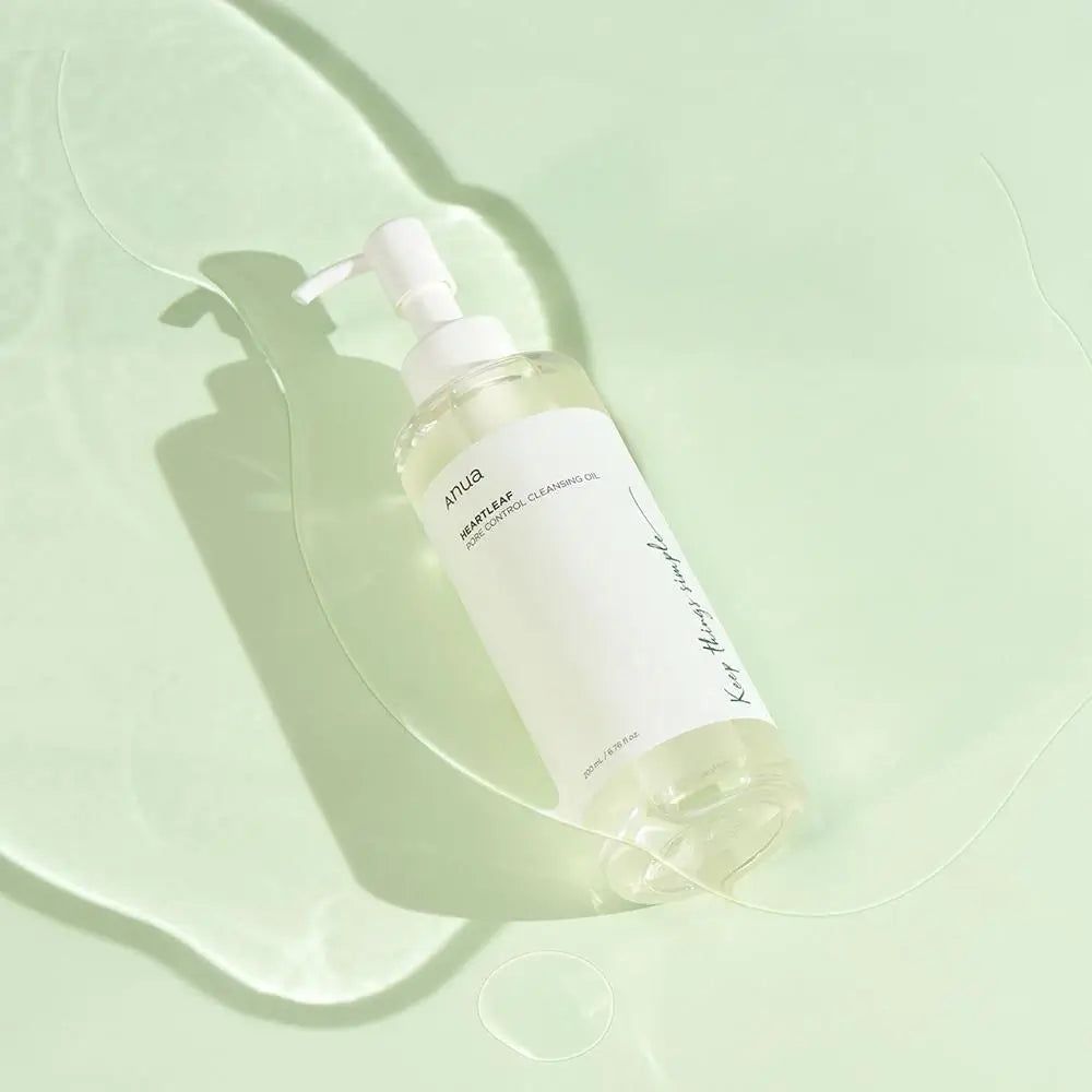 Klare Flasche mit Pumpe. Aufschrift: Anua Heartleaf Pore Control Cleansing Oil. Auf hellem Hintergrund mit Wasser.