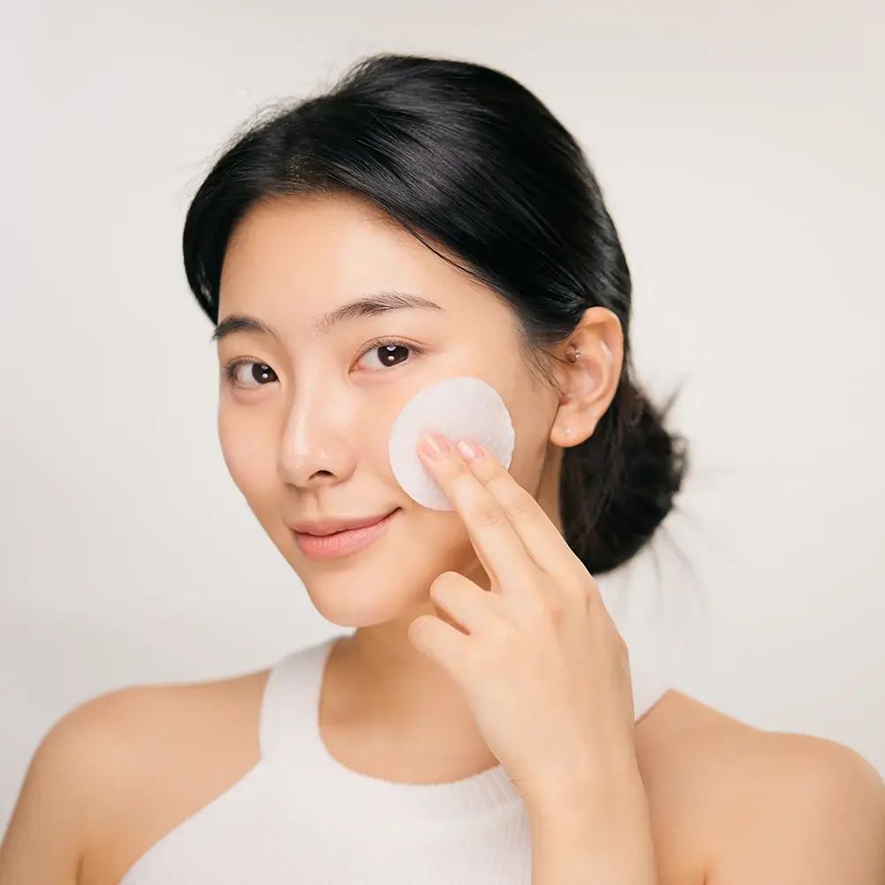 Junge Frau mit einem Wattepad auf dem Gesicht. Sie berührt ihre Haut mit dem Pad. Helles Hintergrund.