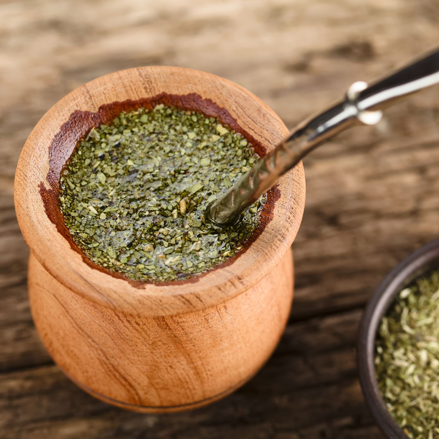 Holzbecher mit Yerba Mate Tee und Metall-Trinkhalm. Daneben eine kleine Schale mit getrockneten Blättern. Auf Holzuntergrund.