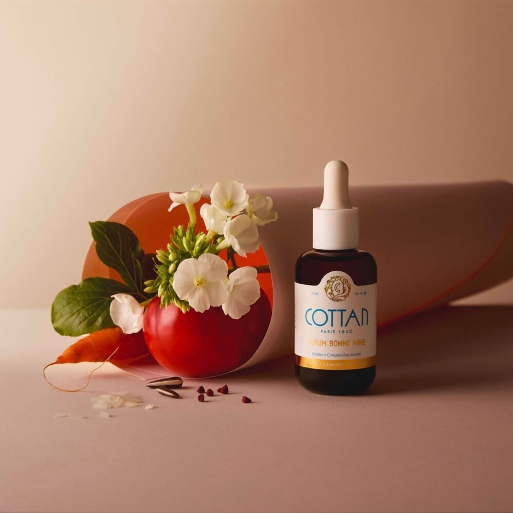 Serumflasche mit Tropfer, daneben eine rote Schale mit Blumen und Gemüse. Aufschrift: COTTAN, Serum Bonne Mine.