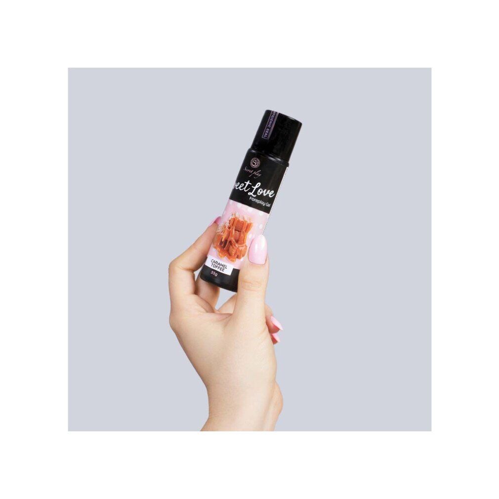 Hand hält eine schwarze Flasche. Aufschrift: Sweet Love Foreplay Gel. Abbildung von Karamell-Toffee. 55g. Rosa Hintergrund.