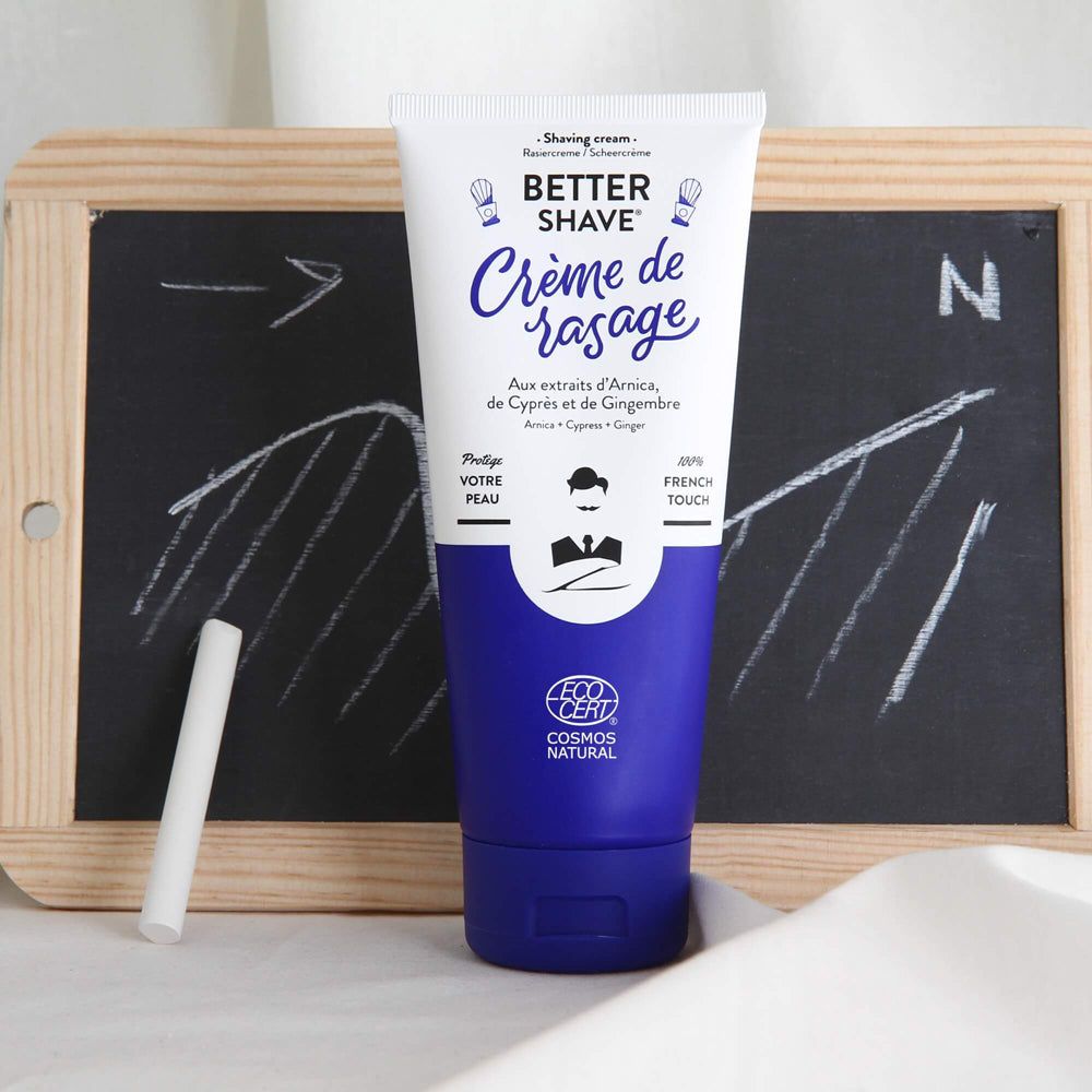 Rasiercreme-Tube vor einer Tafel mit Kreide. Tube mit blauem Boden, weißem Hintergrund und Text. COSMOS NATURAL-Logo.