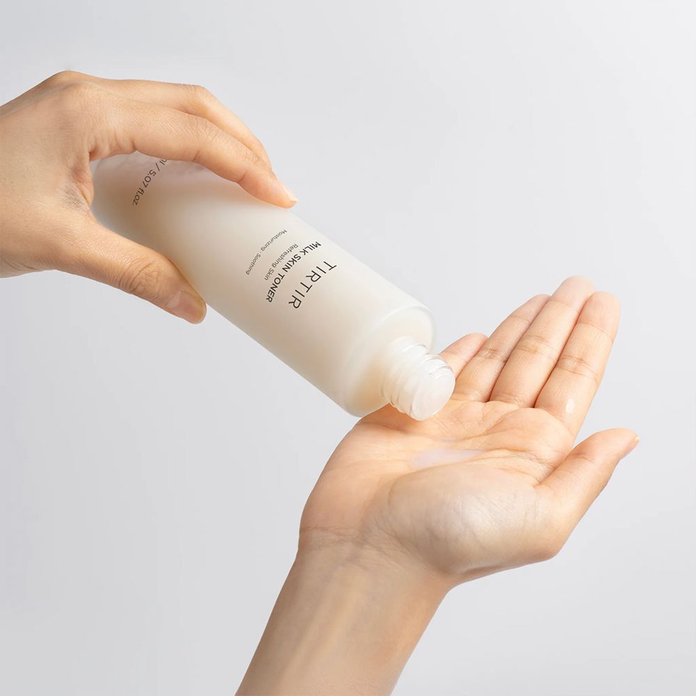 Hände halten eine Flasche TIRTIR Milk Skin Toner. Flüssigkeit wird in die Handfläche gegossen. Flasche mit silbernem Deckel.
