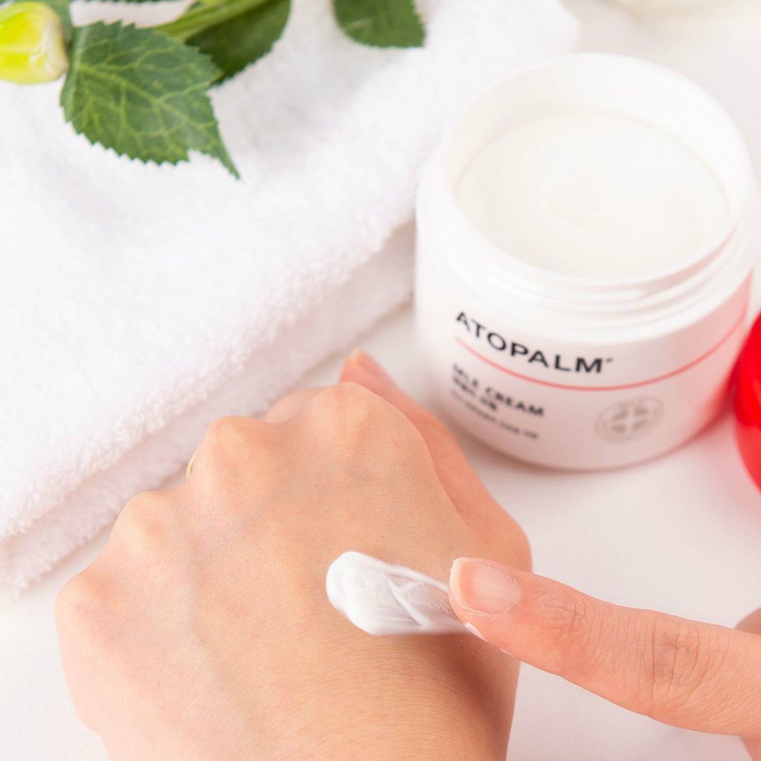 Hand mit Creme auf dem Finger. Offener Tiegel mit Creme daneben. Handtuch und Blüte im Hintergrund.