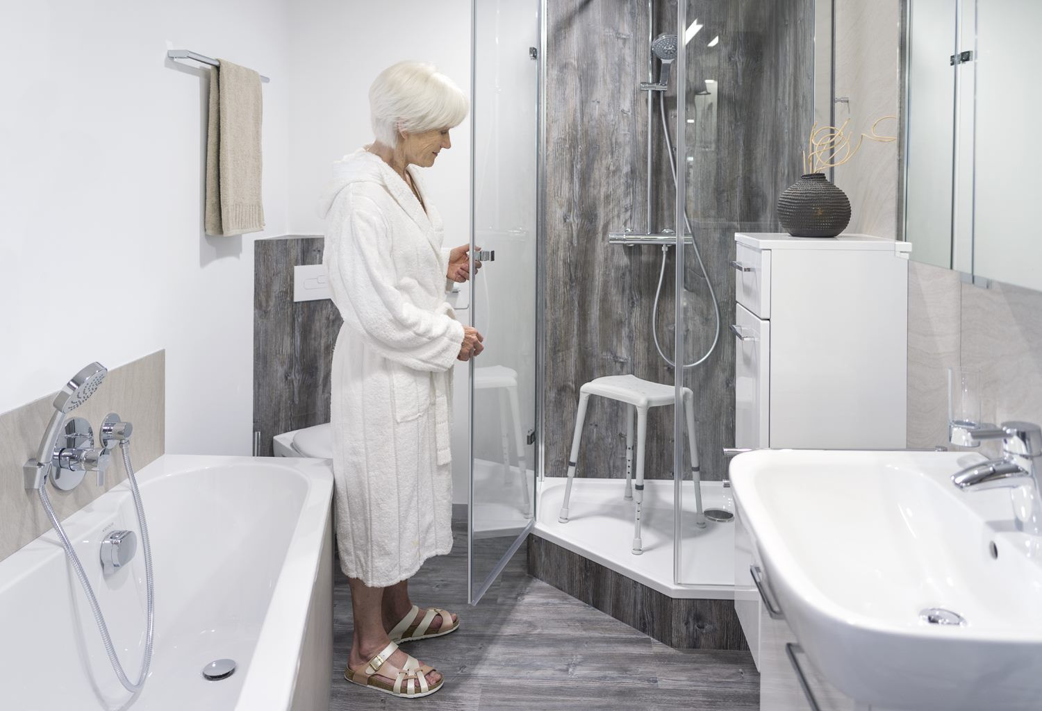 Ältere Frau in einem Badezimmer. Sie steht vor einer Dusche und neben einem Duschhocker. Das Badezimmer ist hell und modern.