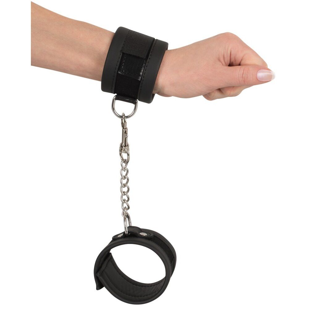 Handfessel am Handgelenk. Schwarzes, gepolstertes Material. Kette mit Karabinerverschluss. Andere Hand frei.