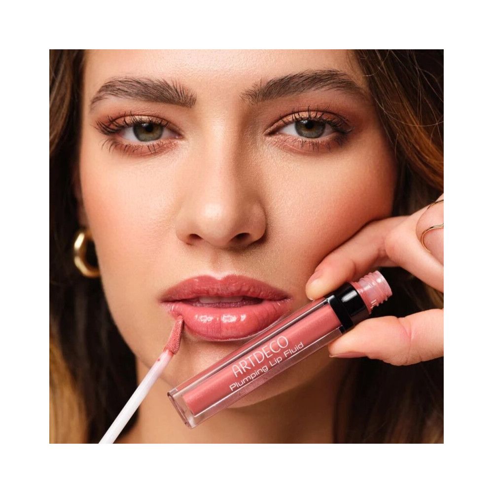 Frau trägt Artdeco Plumping Lip Fluid 028 Goddess auf. Produkt und Applikator sichtbar. Rosa Lippenstift.