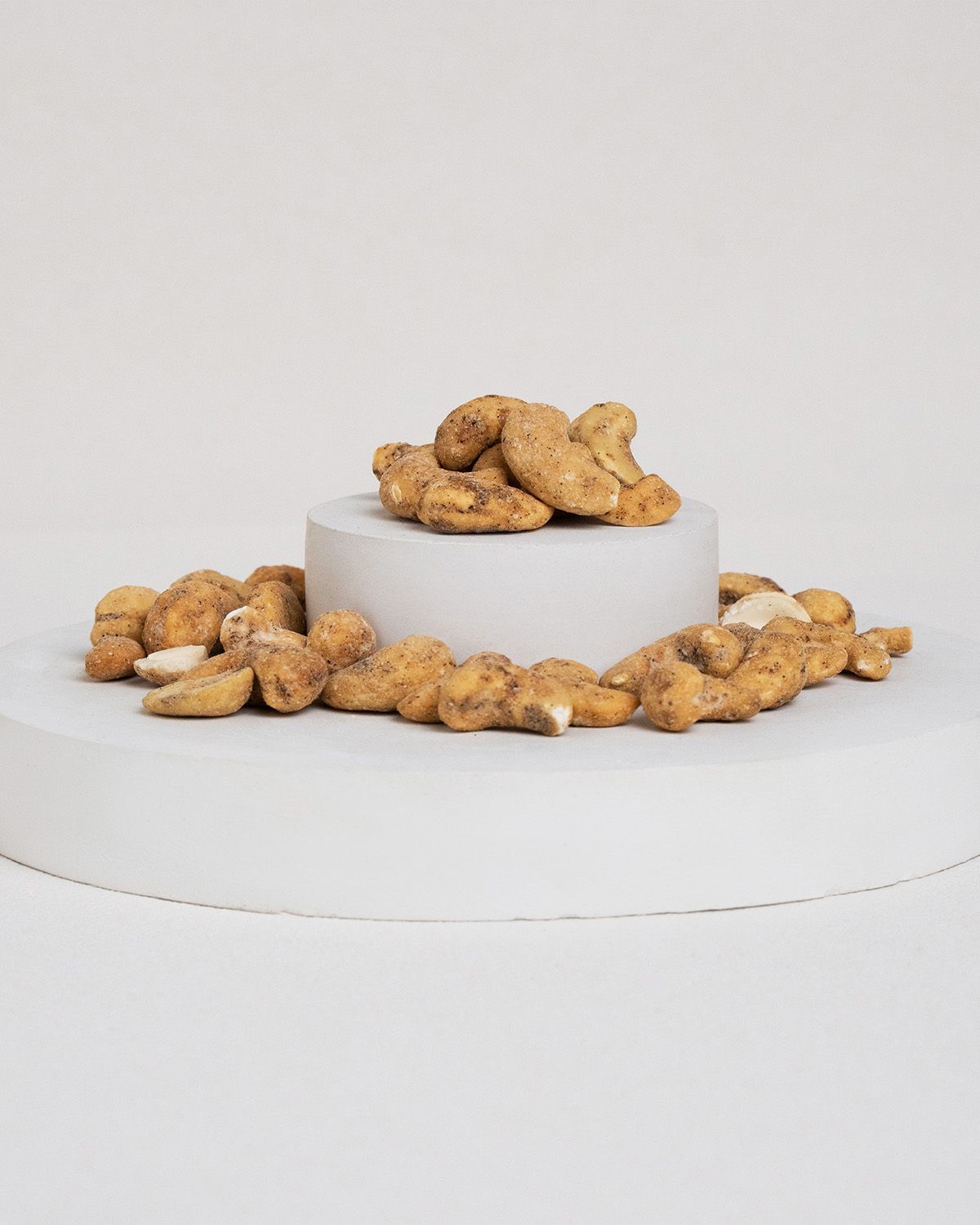 Cashewkerne auf einem weißen Podest. Einige Kerne liegen daneben. Helle, runde Basis.