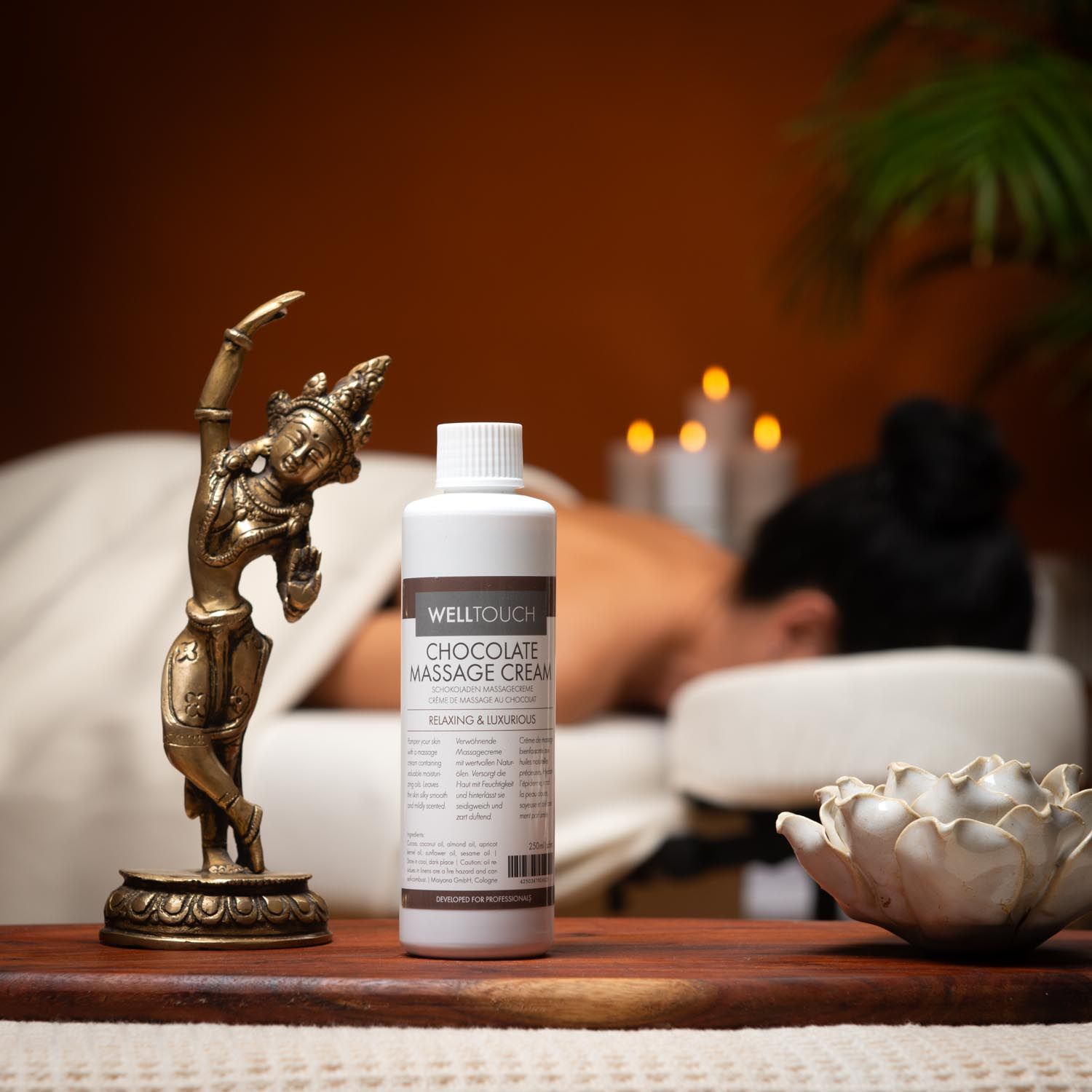 Weiße Flasche mit Welltouch Schokoladenmassage-Creme. Aufschrift: Chocolate Massage Cream. Person im Hintergrund auf Massageliege.