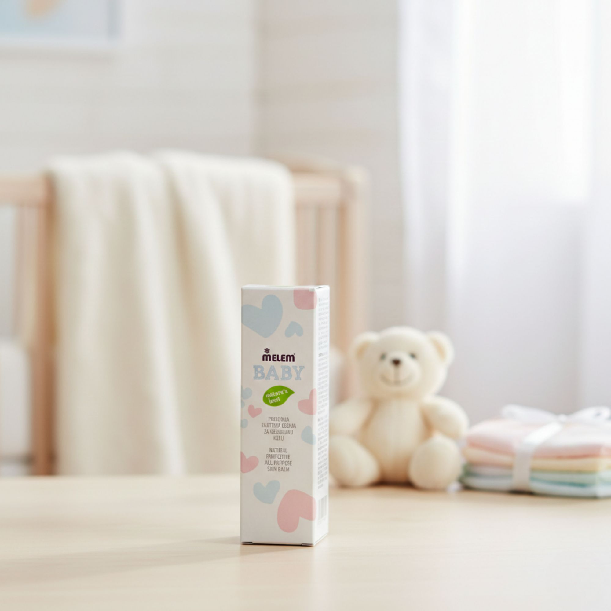 Kartonverpackung "Melem Baby" auf einem Tisch. Text: "Natural Protective All-Purpose Skin Balm". Hintergrund: Babybett, Teddybär.