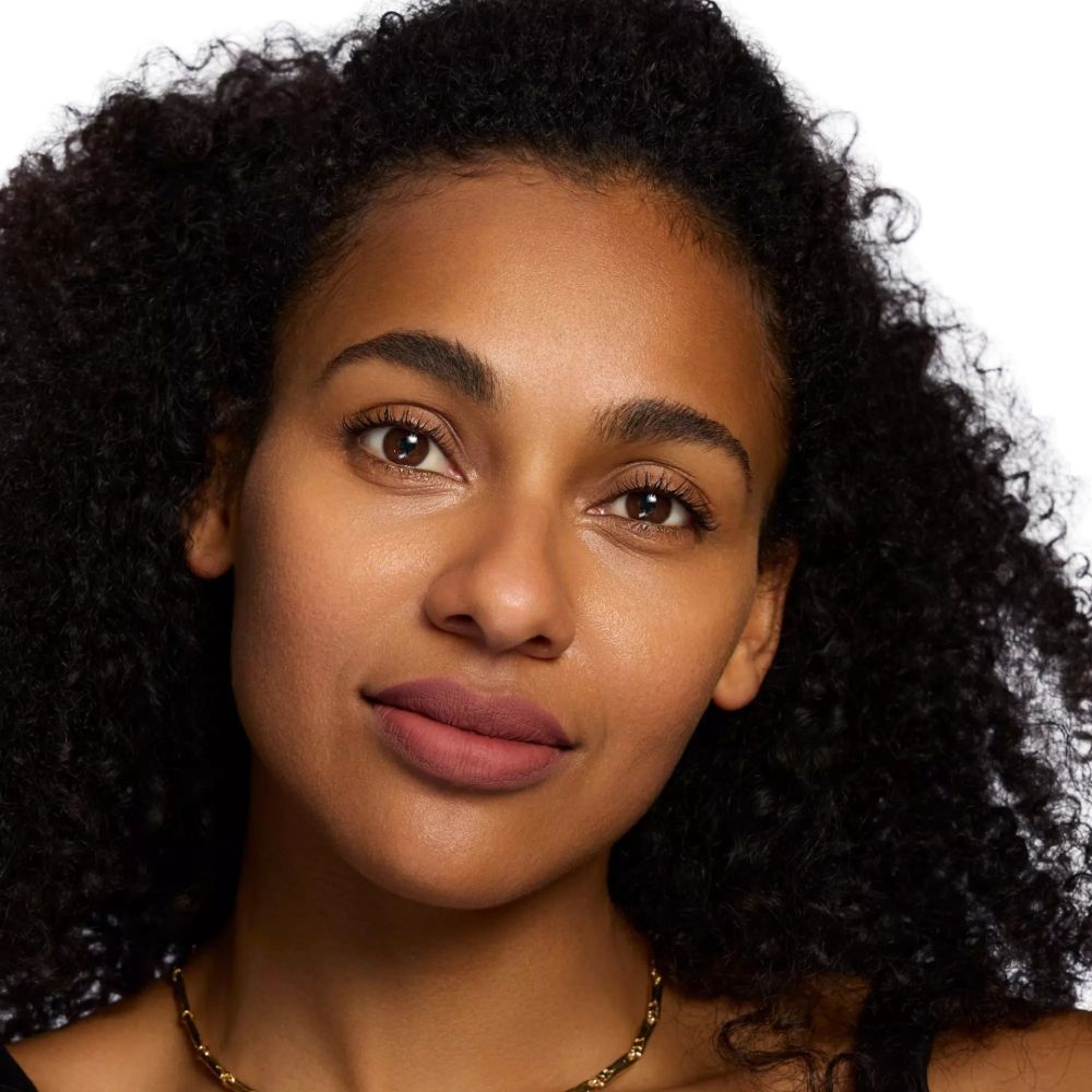 Porträt einer Frau mit Lippenstift. Dunkle Haut, lockiges Haar, goldene Halskette. Lippenstift in Nude-Ton.