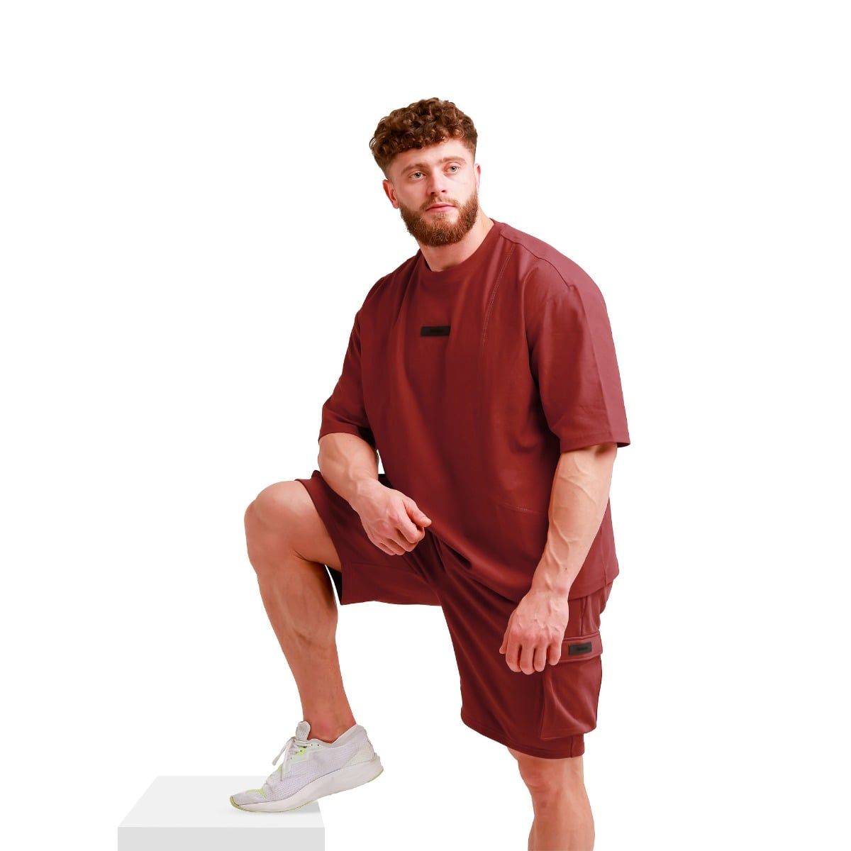 Mann in rotem Oversize T-Shirt und Shorts, steht auf einem weißen Podest, Bein aufgestellt.