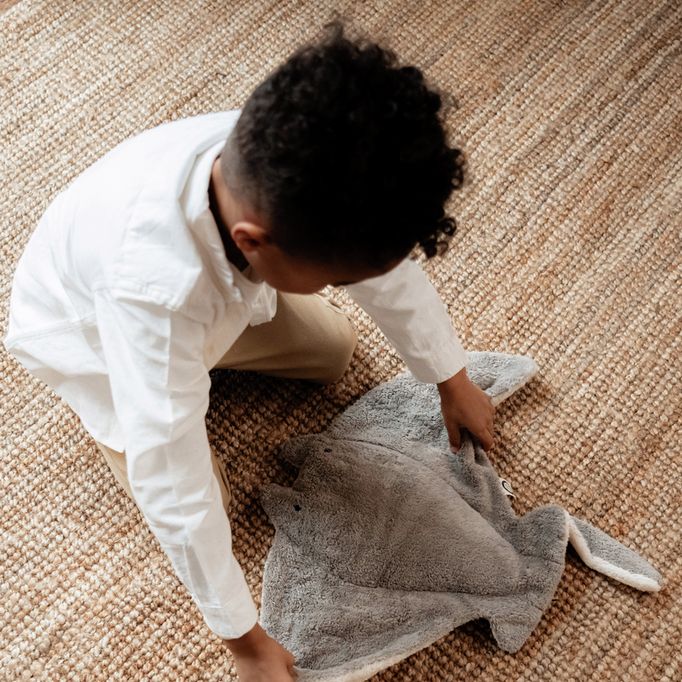Kind beugt sich über ein graues Kuscheltier Manta auf einem Teppich.