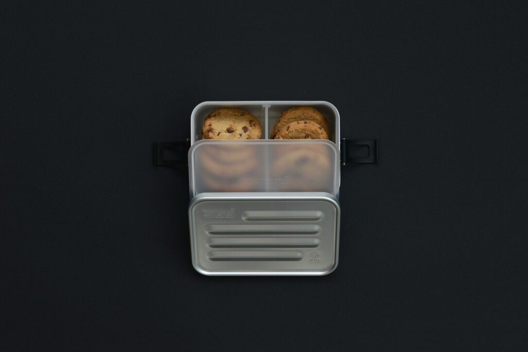 Geöffnete SIGG Lunchbox Metal Box Plus S mit Keksen. Silberfarbenes Metall, schwarze Verschlüsse. Innenaufteilung mit transparentem Einsatz.