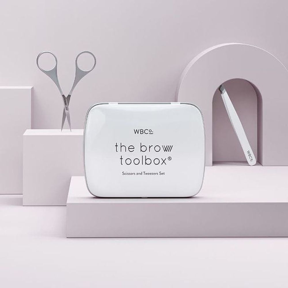 Weißes Etui mit Aufschrift 'the brow toolbox'. Pinzette und Schere daneben. 'WBCo.'-Logo auf dem Etui.