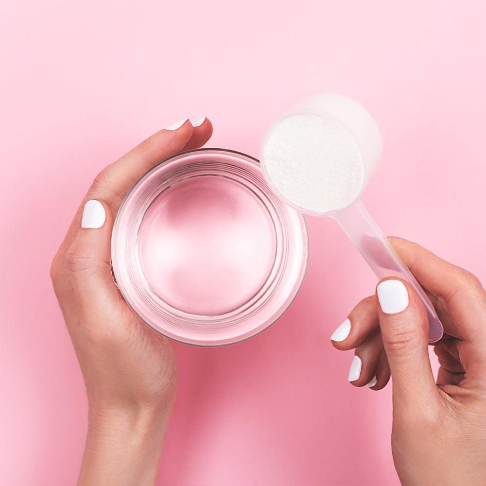 Hände halten Glas mit Flüssigkeit und Messlöffel mit Pulver. Rosa Hintergrund.