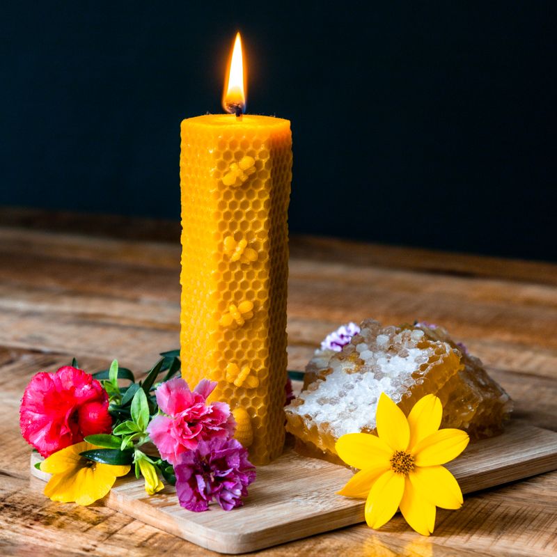 Gelbe Wachskerze mit Bienenwabenmuster, brennend. Neben Honigwaben und Blumen auf Holzbrett.