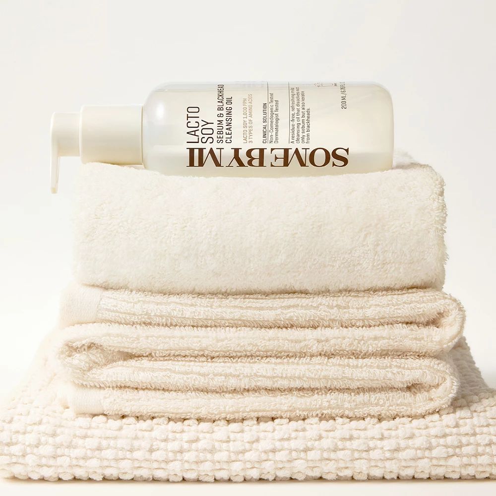Flasche mit Pumpe, gefüllt mit Flüssigkeit, auf einem Stapel Handtücher. Aufschrift: SOMEBYMI Lacto Soy Sebum & Blackhead Cleansing Oil.