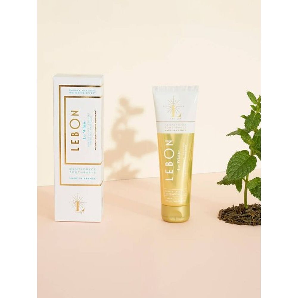 LEBON Le White Zahnpasta-Tube und Schachtel. Beide weiß-goldfarben. Schachtel links, Tube rechts. Minze-Pflanze im Hintergrund.