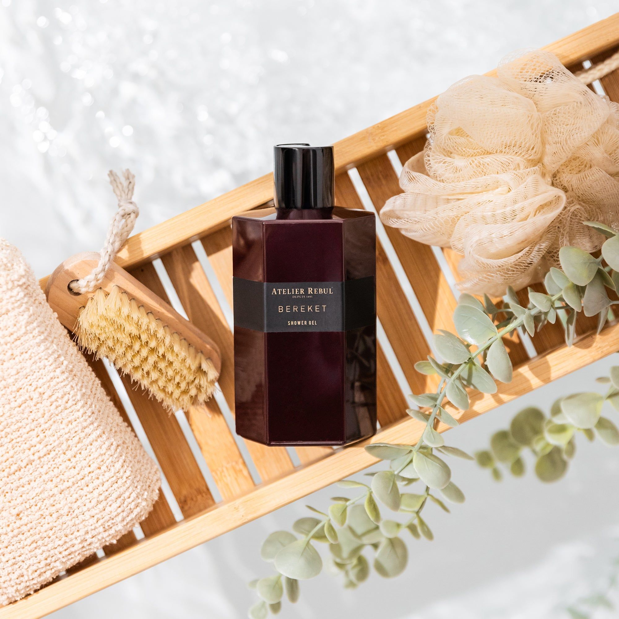 Bordeauxfarbenes Duschgel auf einem Holztablett. Mit Schwamm, Bürste und Zweigen dekoriert. Aufschrift: Atelier Rebul Bereket.
