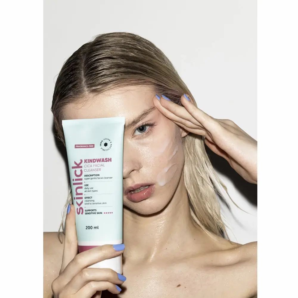 Gesichtsreinigungsgel-Tube vor Gesicht. Hellgrün-weiß, pinkfarbener Streifen. Text: Kindwash, Cica Facial Cleanser, 200 ml. Frau mit Produkt.