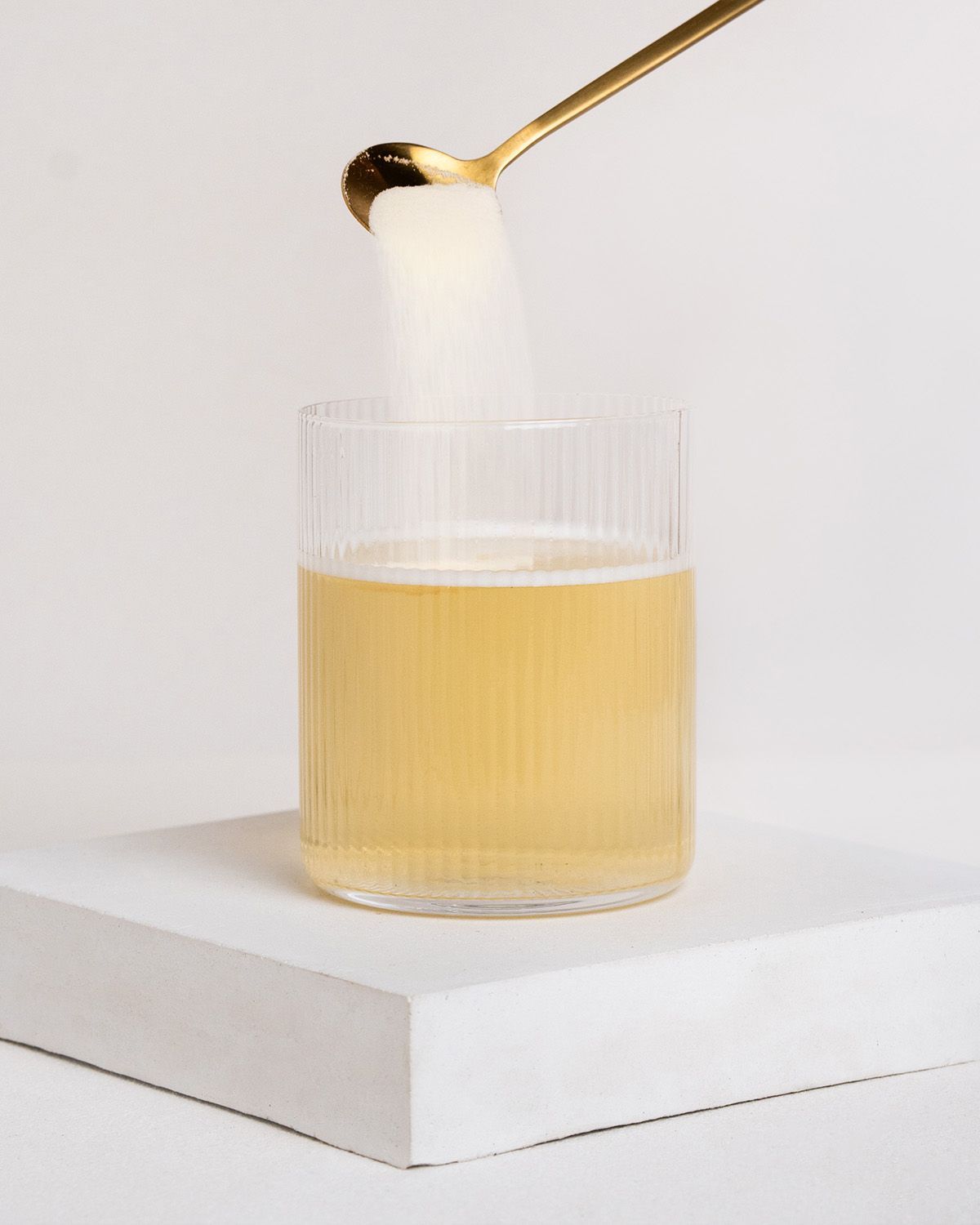 Glas mit Flüssigkeit, goldener Löffel mit Pulver. Pulver wird in das Glas gegeben. Geriffeltes Glas auf weißem Podest.