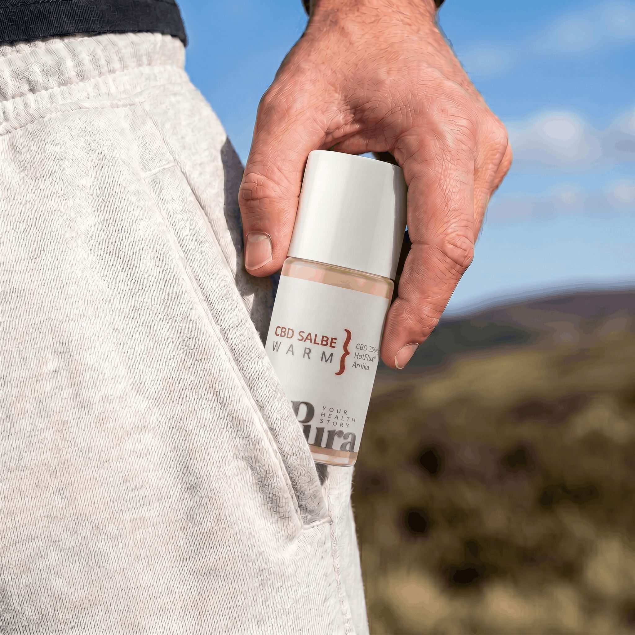 Hand steckt eine CBD Salbe Warm Roll-On Flasche in die Hosentasche. Hintergrund ist eine Landschaft.