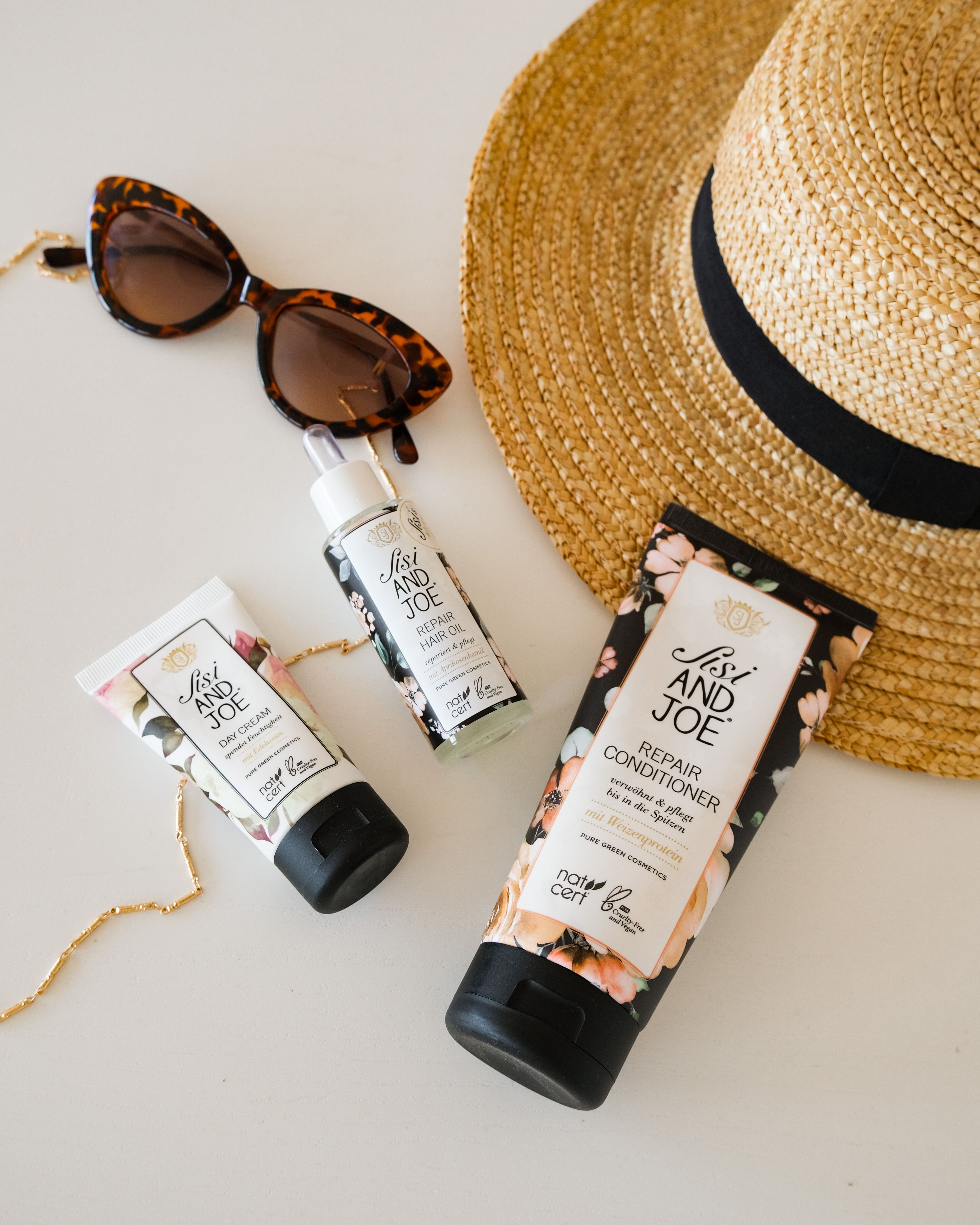 Sonnenbrille, Sonnenhut und drei Kosmetikprodukte mit floralem Muster. Handöl, Repair Conditioner und Day Cream.