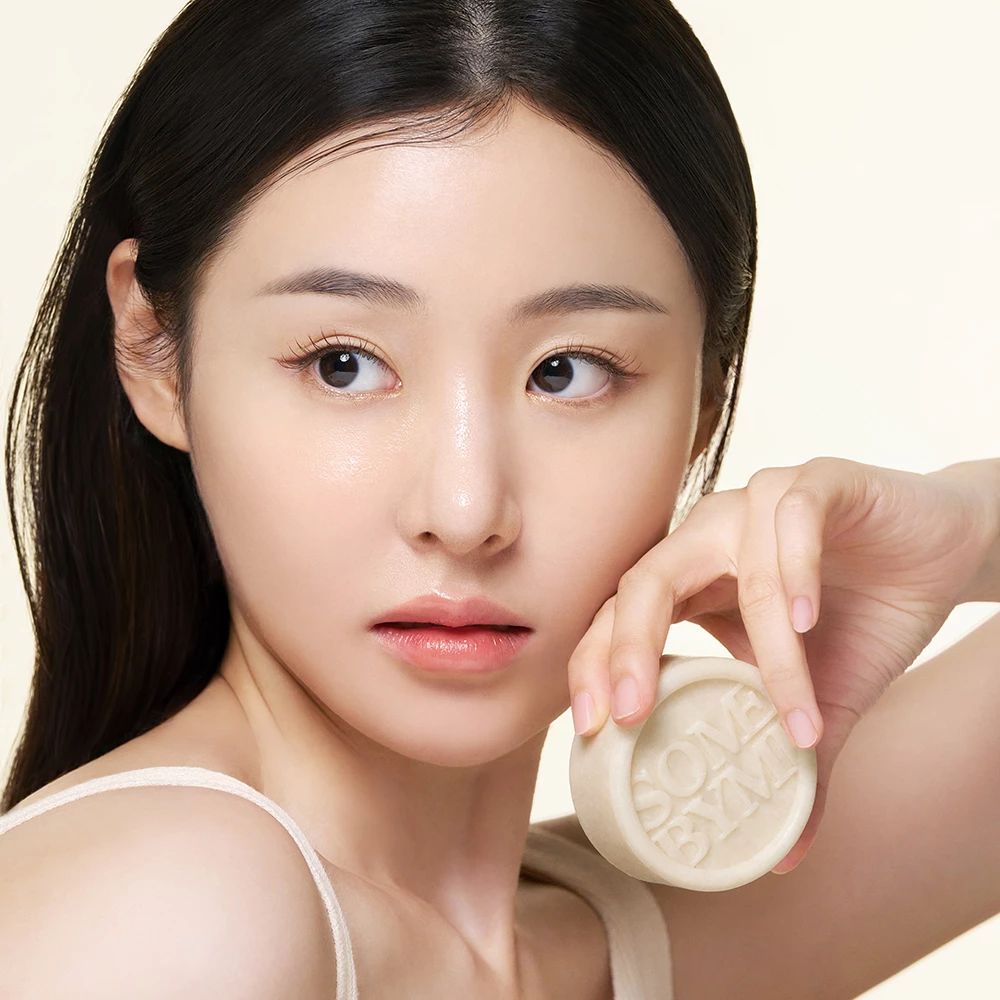 Eine Frau hält eine runde Seife mit dem Text SOME BYMI. Sie berührt ihr Gesicht.