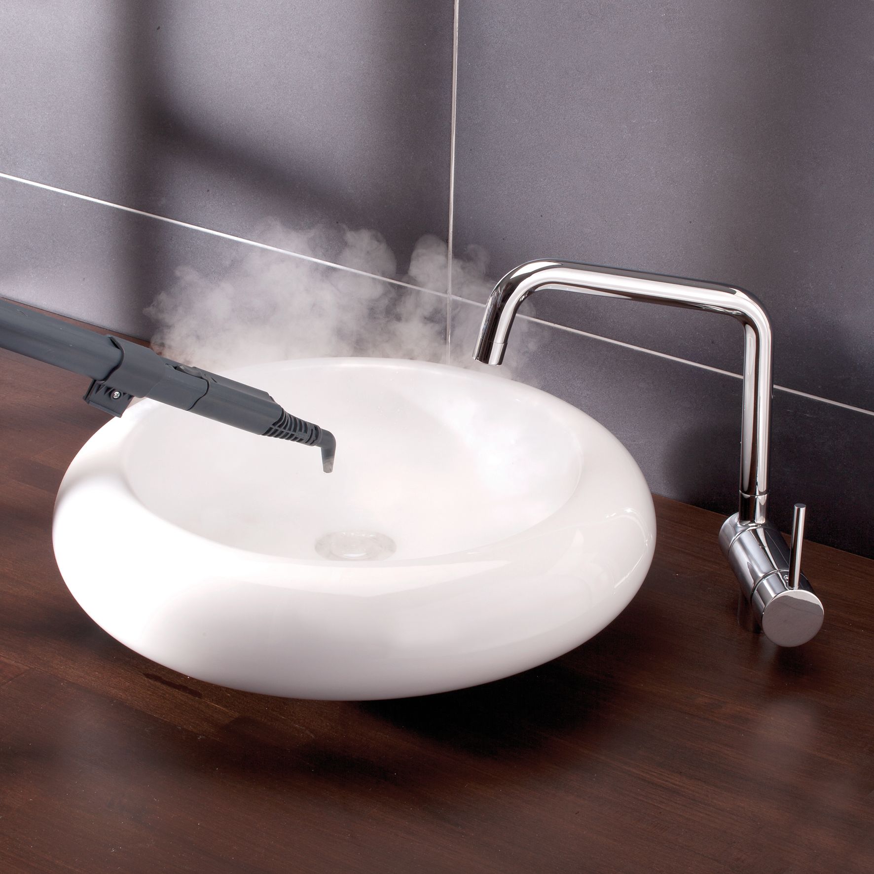 Dampfstrahl aus einer Düse in ein Waschbecken. Waschbecken weiß, rund. Wasserhahn aus Metall. Hintergrund: Fliesenwand.